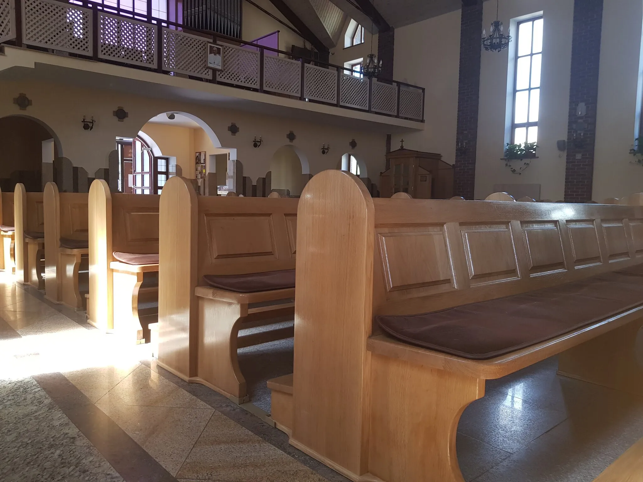 Rzędy jasnych drewnianych ławek w nawie kościoła, w tle balustrada chóru