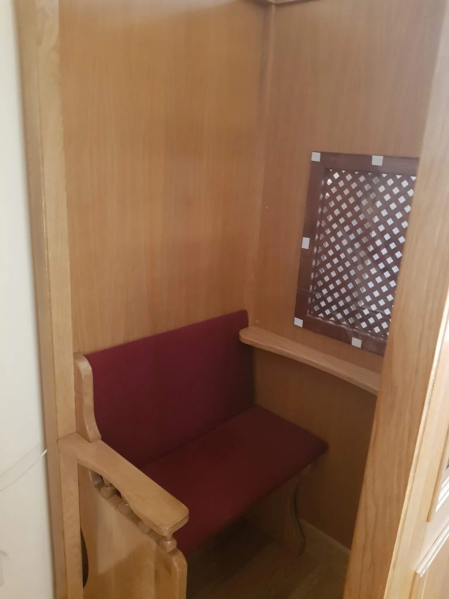 Wnętrze konfesjonału z ławką w bordowym obiciu i kratą w ściance działowej